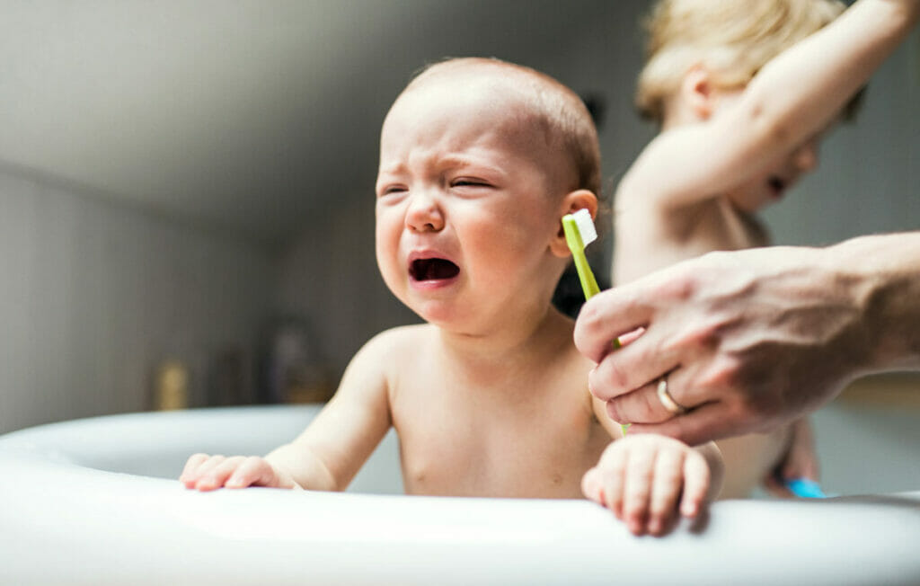 Barn i badekar som gråter, etter å ha fått sin første tann. Forelder som holder en tannbørste for å gi barnet sin første tannpuss.